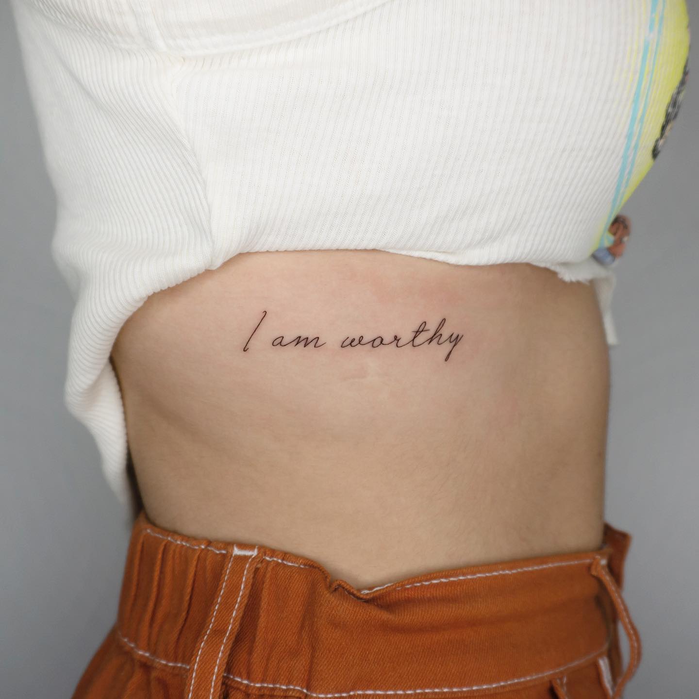 Script "I am worthy" self love tattoo along the hip and lower ribs