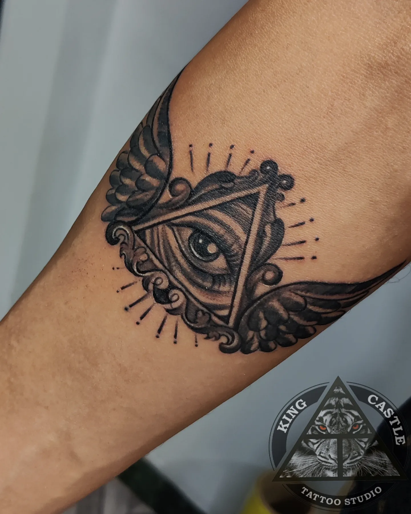Winged third eye tattoo on the inner forearm, the eye set inside a triangle with feathered wings and radiating black and grey rays.