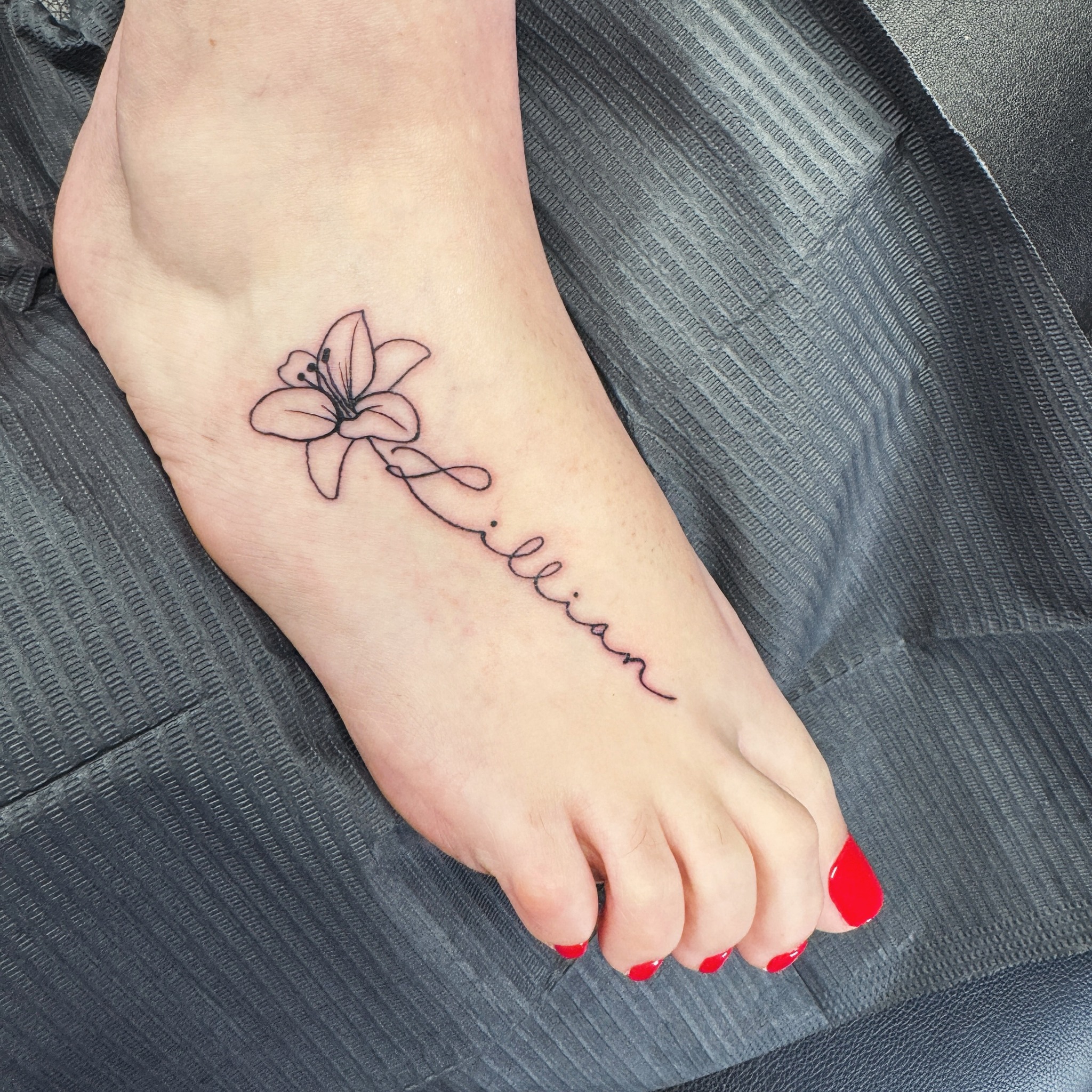 Name tattoo on the foot wrapped around a fine line lily outline