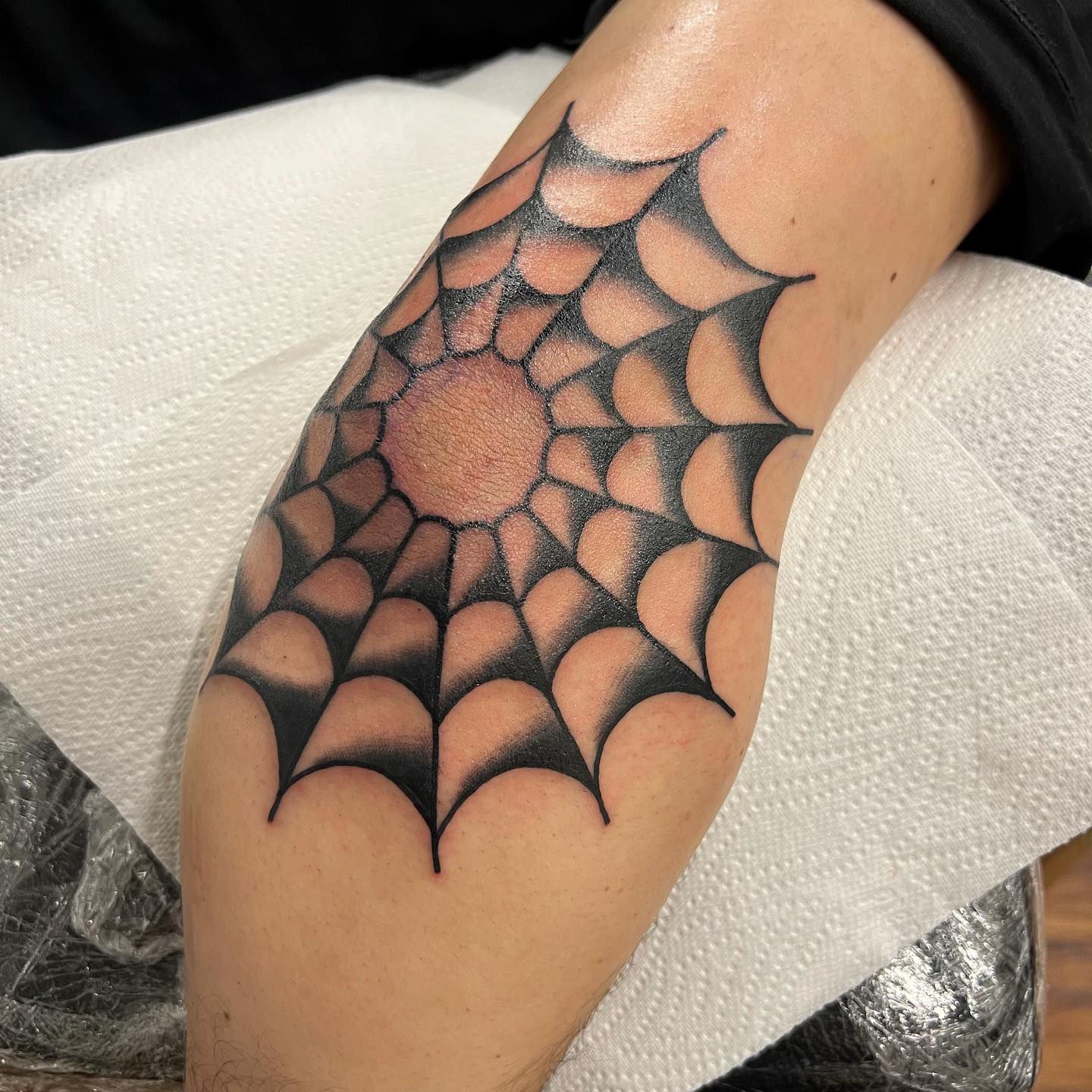 Black spider web tattoo radiating across the outer elbow, an elbow tattoo design built around the joint as its centre point.