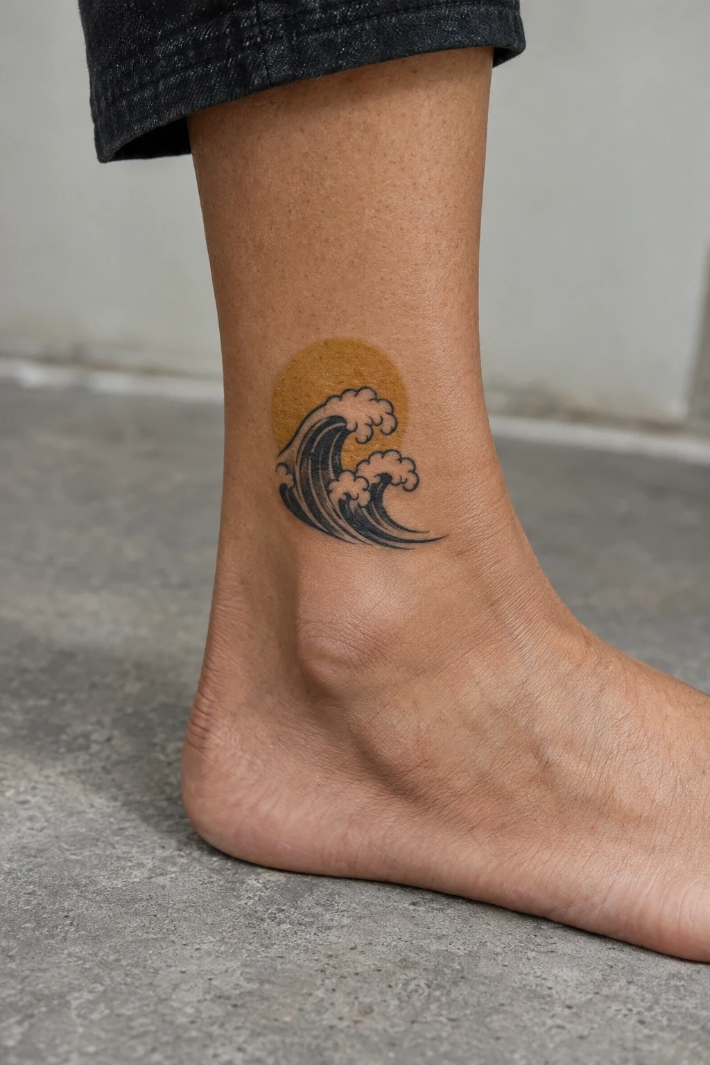 Black wave tattoo with open-skin foam and an ochre sun disk above an ankle